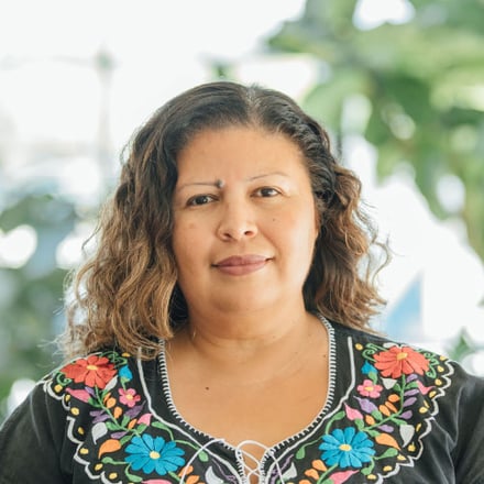 A woman, Rosa Cabrera, poses for a headshot with blurry foliage in the background. 