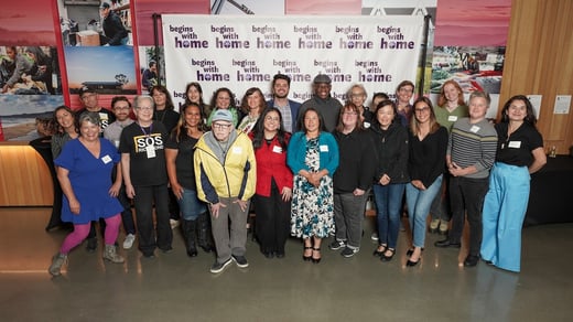 About two dozen people stand together and smile in front of a backdrop that says, “Begins with Home.”