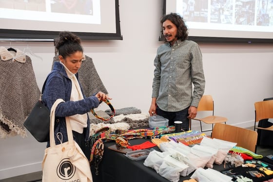 A person shops and speaks to a business owner at a table display with handmade jewelry and accessories.