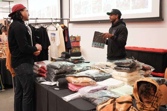 Two men talk over a table display of t-shirts.