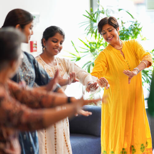A group of women in traditional attire dance together, with one woman in a yellow dress leading and smiling.