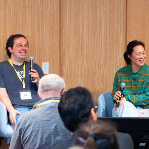 Two people are seated on a panel, both holding microphones and smiling; the man on the left, Boris Power, is wearing a T-shirt with a logo and a lanyard, while the woman on the right, Priscilla Chan, is wearing a green striped shirt. An audience is visible in the foreground.
