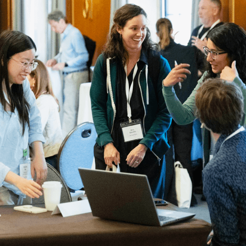 A group of people gathered around a table with a laptop, engaging in a lively discussion and smiling, at a conference or workshop.