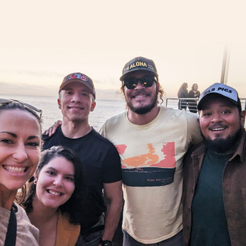 A group of five people are smiling and posing for a selfie at an outdoor location with the ocean in the background, wearing casual clothing, including hats and sunglasses.