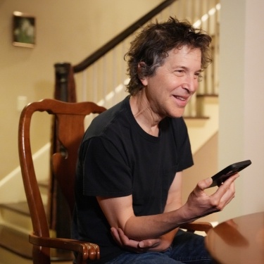 David Baker in a black t-shirt, sitting in a dining chair, smiling with his cell phone in his hand.