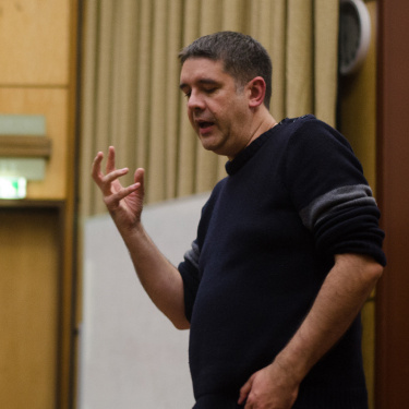 Dan Quine in a dark sweater and jeans gestures while speaking in a lecture hall.