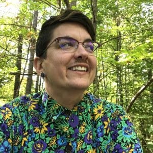 Headshot of Alex Fischer, wearing a bright flowery shirt in a forest, is looking up and to the right while smiling.