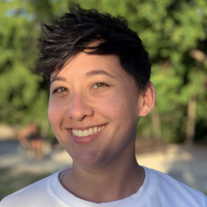 Headshot of Annie Sullivan-Chin, wearing a white shirt with blurred trees in the background, looking directly at the camera and smiling.