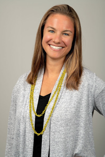 A professional headshot of a smiling woman with light brown hair, wearing a black top layered with a light gray cardigan and accessorized with a long yellow beaded necklace. She stands against a neutral gray background, looking directly at the camera with a warm and confident expression.