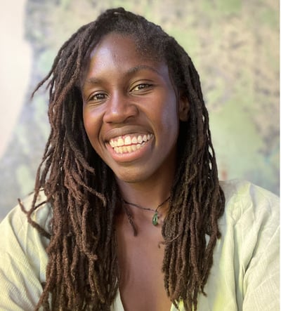 A Black Woman with dreadlocks smiling at the camera. She is wearing a green top with an offset background.