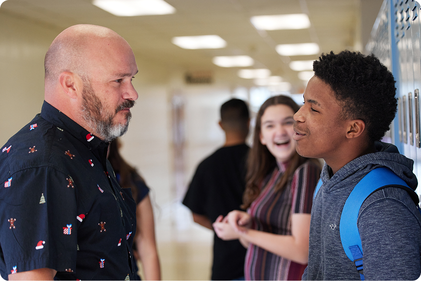 An adult speaks with a student in a school hallway. Two other students stand in the background.