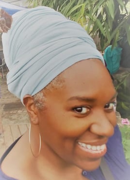 Black woman in a purple blouse wearing a large hoop earring. She is smiling and wearing a teal headwrap
