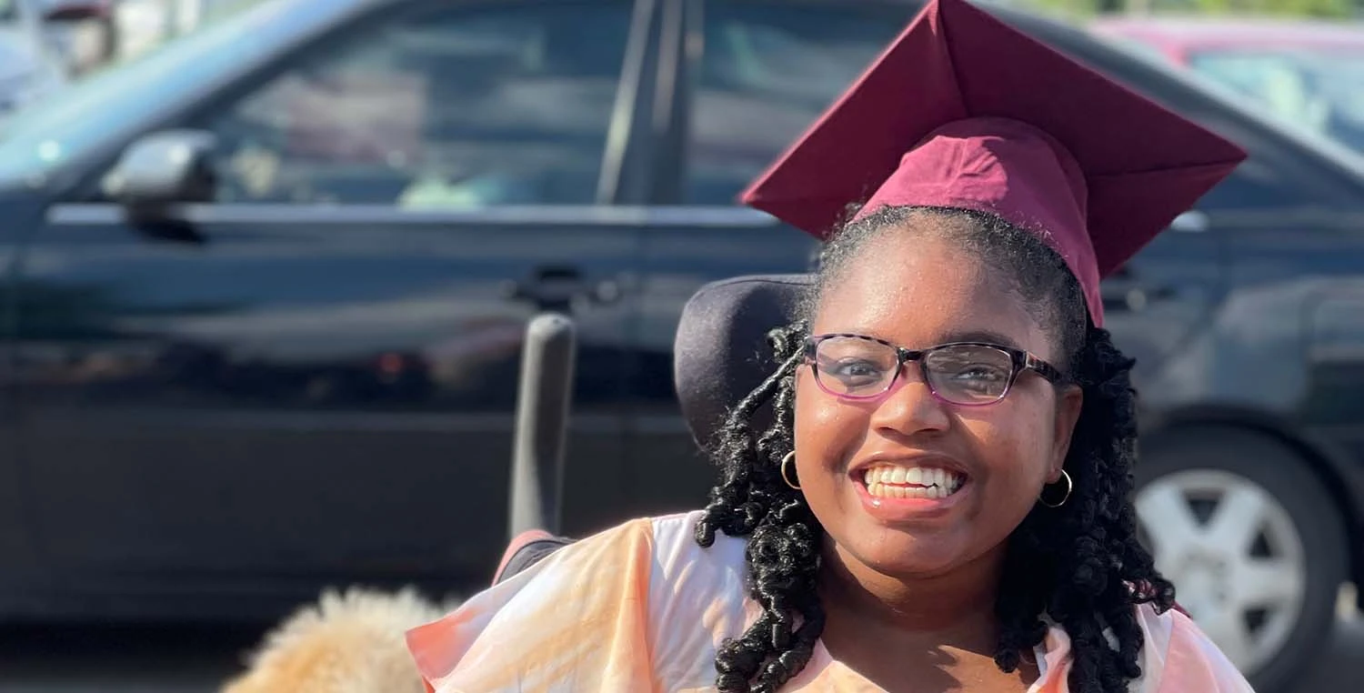 Khari McCrary, a teenage girl, wearing a graduation cap and sitting in a wheelchair smiles in the school parking lot.